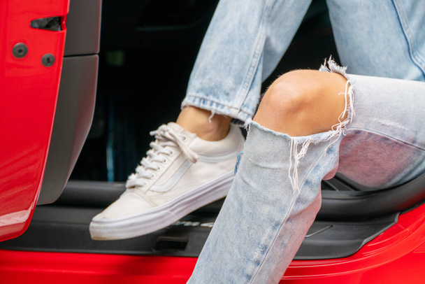 A close-up shot of a girl sitting in a red car wearing blue torn jeans and white shoes - Foto, Imagem