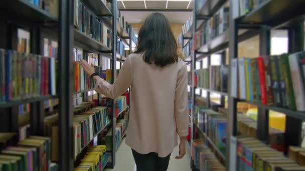 Achteraanzicht van tienermeisje met rugzak lopend in schoolbibliotheek. Tiener kind loopt tussen planken in de boekenwinkel. Onderwijs, leefstijl van studenten en lesconcept. - Video