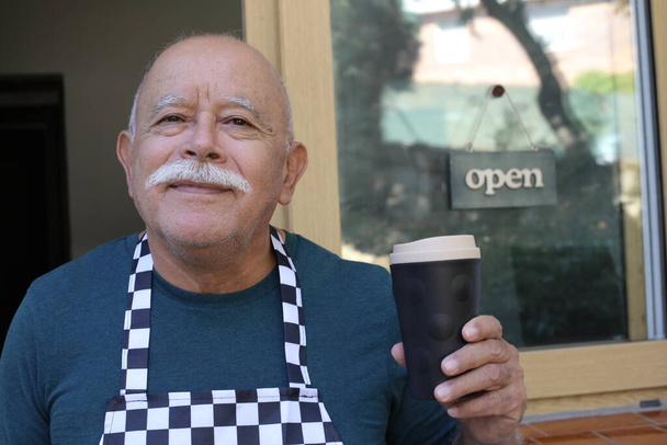 γκρο πλαν πορτρέτο του όμορφου πρεσβύτερου barista κρατώντας χάρτινο φλιτζάνι καφέ μπροστά από γυάλινη πόρτα του καφέ - Φωτογραφία, εικόνα