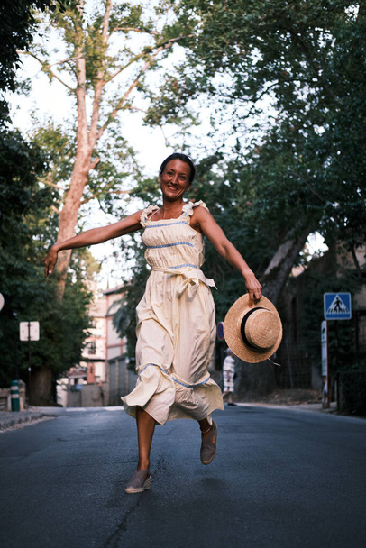Молодая женщина в платье и шляпе бегает и прыгает по дороге в окружении деревьев - Фото, изображение