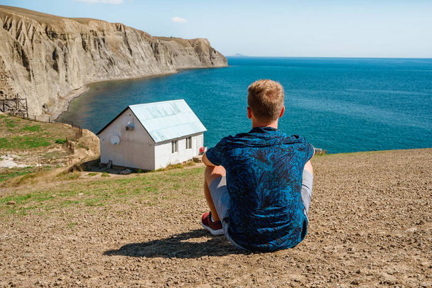 Вид сзади на молодого человека перед белым одиноким домом на краю скалы с живописным горным ландшафтом и видом на море - Фото, изображение