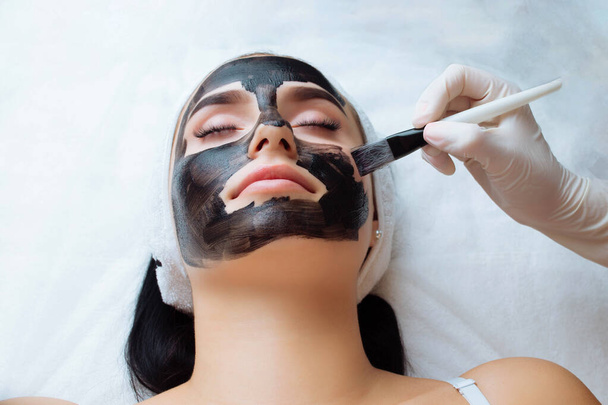 cosmetoloog toepassen van zwart masker op mooie vrouw gezicht dragen van zwarte handschoenen, prachtige vrouw in spa met gezichtsbehandelingen. - Foto, afbeelding