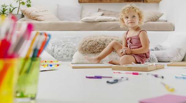 smiling child, beautiful little girl with blue eyes and curly blonde hair, draws and writes cheerfully playing with coloured chalks, preschool learning activity at home, concept of healthy growing - Photo, Image