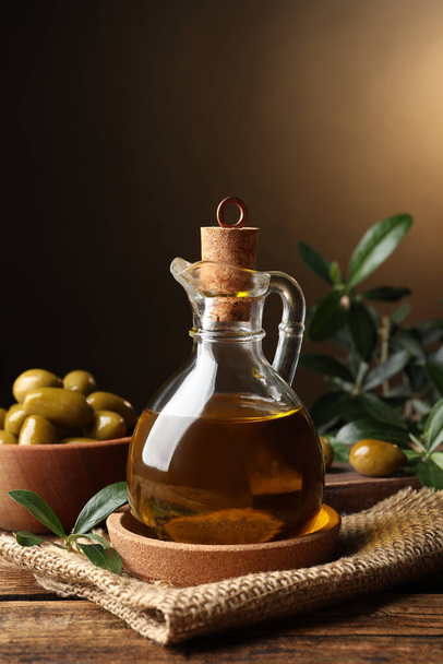 Skleněný džbánek s olejem, zralými olivami a zelenými listy na dřevěném stole - Fotografie, Obrázek
