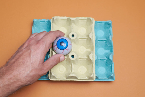 Ojo azul en un cartón de huevo con fondo naranja, ojo ensangrentado en la mano - Foto, Imagen