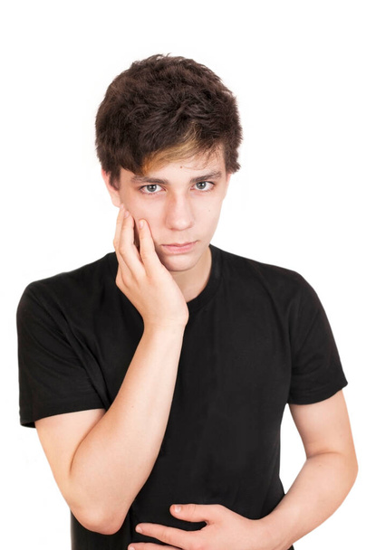 Ένας έφηβος με θλιμμένο πρόσωπο σε λευκό φόντο με μαύρο μπλουζάκι κρατάει το μάγουλό του και λυπάται για τον πονόδοντο. Κάθετη φωτογραφία. Ήρθε η ώρα να πάει στον οδοντίατρο. - Φωτογραφία, εικόνα