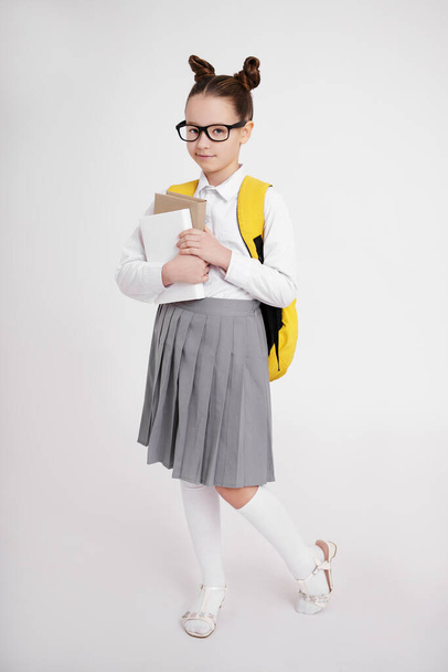 full length portrait of cute school girl in school uniform and eyeglasses with backpack holding books over white background - 写真・画像