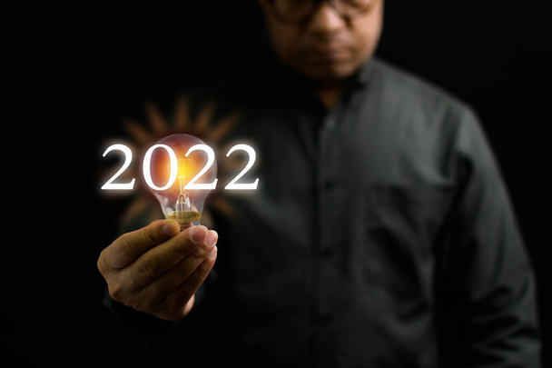 Close-up, A man holding a light bulb is the concept of the startup that drives global business. Bright lights give hope to small businesses. 2022 New Year concept, Copy space, Black Blurred background - Photo, Image