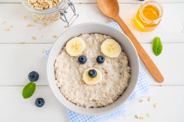 Funny bowl with oat porridge with bear faces made of fruits and berries on a white wooden background. Food for kids idea, top view, copy space. - Photo, Image