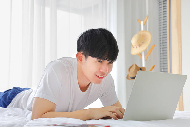 Jeune homme asiatique indépendant travaillant à partir de la maison en utilisant un ordinateur portable tout en étant assis sur le lit avec espace de copie - Photo, image