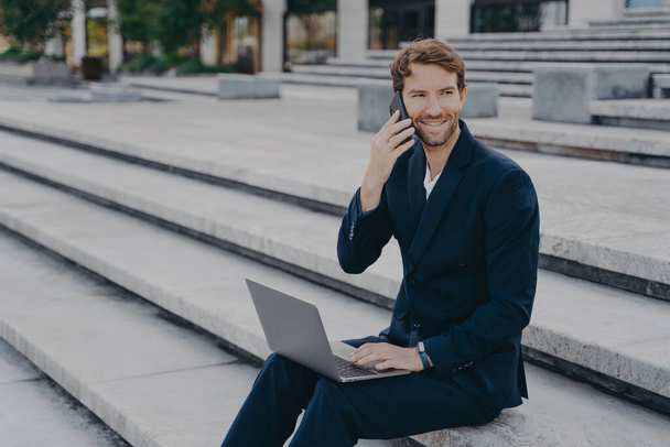 Jonge aantrekkelijke zakenman werkt op afstand op laptop, spreken op smartphone buiten - Foto, afbeelding