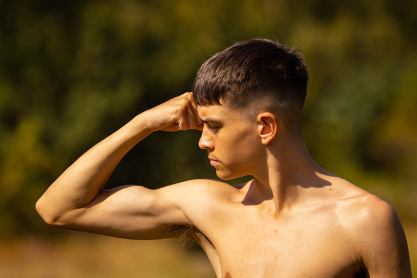 An eighteen year old teenage boy flexing his muscles in a park on a warm summer's day while shirtless - 写真・画像