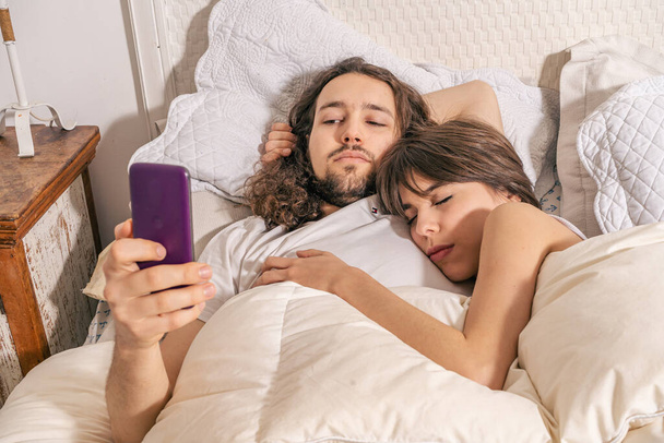 glückliches Paar, das sich nach dem Aufwachen im Bett auf dem Handy unterhält. Mann lächelt und schreibt SMS, seine Freundin checkt soziale Netzwerke. Online-Kommunikation, mobile Anwendungen, Lebensmittelbestellung über das Internet - Foto, Bild