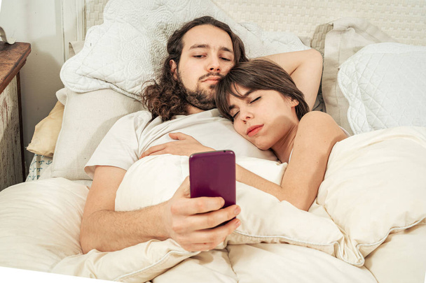 Счастливая пара болтает по мобильному после того, как проснулась лежащей в постели. Мужчина улыбается и пишет, его девушка проверяет социальные сети. Онлайн связь, мобильные приложения, заказ продуктов питания через интернет
 - Фото, изображение