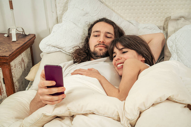 幸せなカップルがベッドに横たわって目を覚ます後携帯でおしゃべり。男は笑みを浮かべて、テキスト メッセージ、ソーシャル ネットワークをチェック彼のガール フレンド。オンラインでのコミュニケーション、モバイル アプリケーション、インターネット経由で食べ物を注文 - 写真・画像