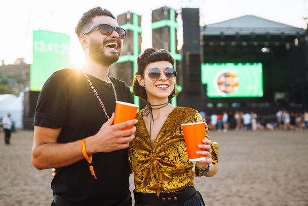 Stylish couple enjoying a performance at a festival together. Young people drinking beer and having fun at music festival at the beach. Youth, party, vacation concept. - Photo, image