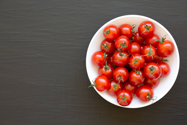Świeże czerwone organiczne pomidory wiśniowe w misce na czarnym tle, widok z góry. Przestrzeń kopiowania. - Zdjęcie, obraz