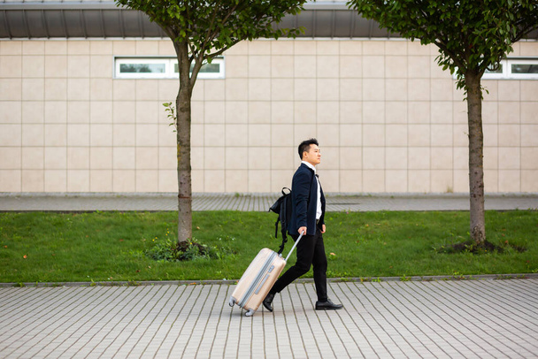 Successful asian businessman near airport and bus station goes with suitcases serious tourist looking at camera - 写真・画像