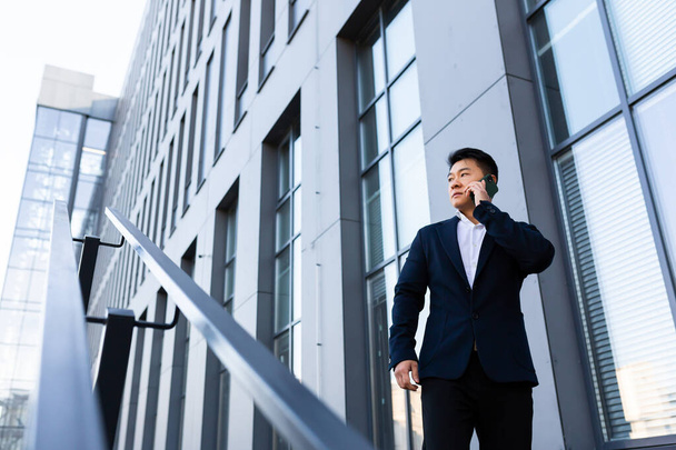 Úspěšný seriózní asijský podnikatel mluví po telefonu v blízkosti kancelářského centra - Fotografie, Obrázek