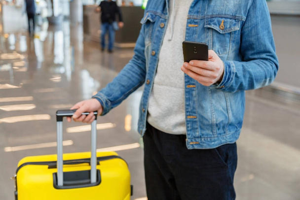  Man using mobile phone at airport. Urban traveler wearing casual holds smartphone, guy texting message while holding suitcase luggage in airport lobby indoor interior after flight. - Photo, Image