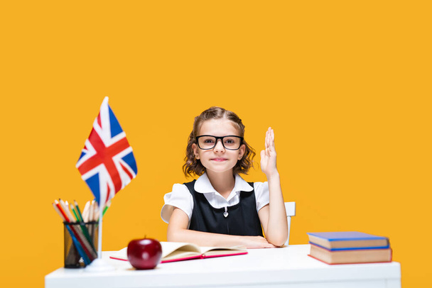 Smiling caucasian schoolgirl raising hand sitting at the desk. English lesson. Great Britain flag - Fotografie, Obrázek