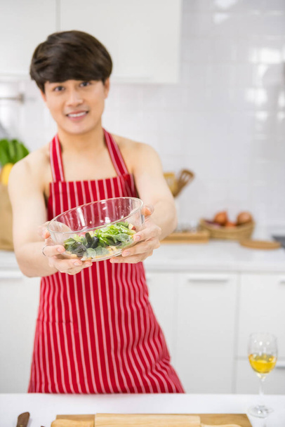 Kırmızı önlüklü seksi LGBT gay genç Asyalı adam mutfakta yemek pişirdikten sonra taze salata tutuyor. Nake gülen adam partneri için sağlıklı yemek pişirir. Hoş homoseksüel, hafta sonu aynı seks ailesi.. - Fotoğraf, Görsel
