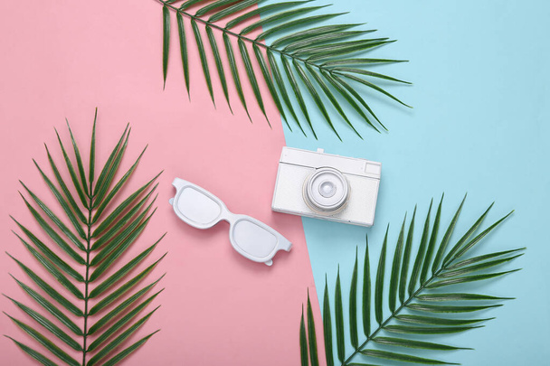 Retro camera, zonnebril en palmbladeren op roze blauwe achtergrond. Tropisch, minimale indeling. Bovenaanzicht. Vlakke plaat - Foto, afbeelding