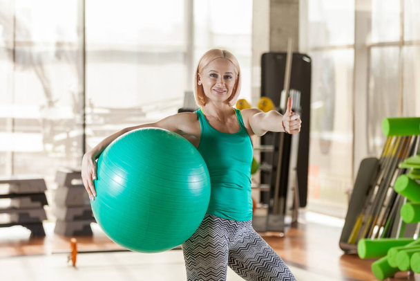 Молодая счастливая фитнес-женщина позирует с фитболом в спортзале. Показывает большие пальцы вверх. Здоровый образ жизни - Фото, изображение