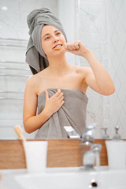 Woman after a shower in a gray towel brushes her teeth with a wooden brush. - Photo, Image
