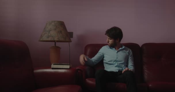 Komea nuori mies istuu sohvalla ja sytyttää pöytälampun - Materiaali, video