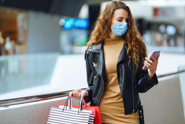Stylish woman in protective medical mask with shopping bags using her phone. Autumn shopping. Fashion, style, Black friday, sale, consumerism concept. - Fotó, kép