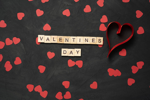 黒いセメントの床に紙の心を持つバレンタインデーのテキストワード木製のブロック。バレンタインデー2月14日の休日。最小限のコンセプトトップビュー. - 写真・画像