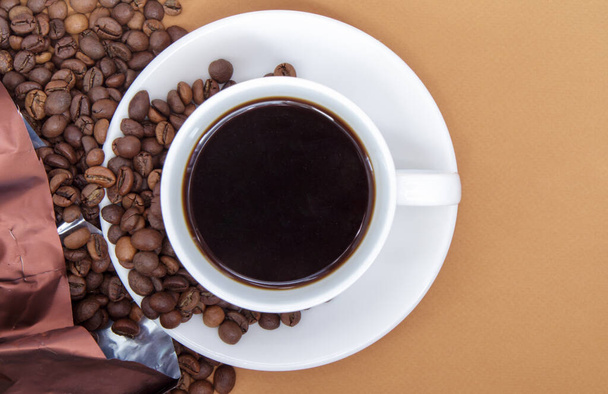 Λευκό φλιτζάνι αμερικάνικου μαύρου καφέ χωρίς γάλα με ένα μάτσο καβουρδισμένους κόκκους καφέ σκορπισμένους σε χάρτινη συσκευασία. Καφές φόντο, πάνω όψη με αντίγραφο χώρο για το λογότυπο ή κείμενο - Φωτογραφία, εικόνα