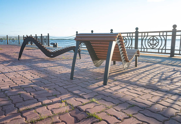 Деревянные скамейки для расслабления необычной формы для отдыха на набережной, на фоне заходящего солнца и голубого неба - Фото, изображение
