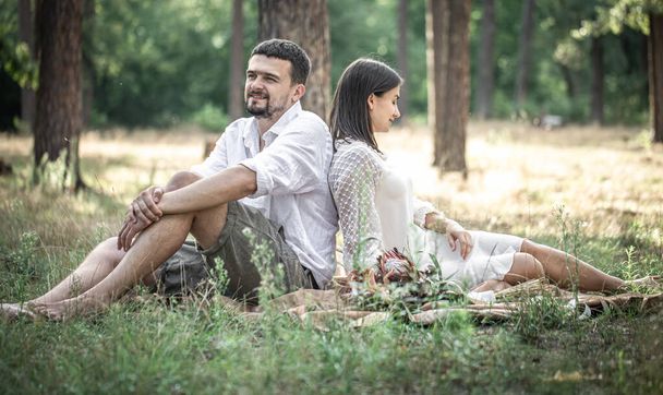 Μια νεαρή γυναίκα με λευκό φόρεμα και ένας άντρας με πουκάμισο κάθονται στο δάσος στο γρασίδι, μια ημερομηνία στη φύση, ειδύλλιο στο γάμο. - Φωτογραφία, εικόνα