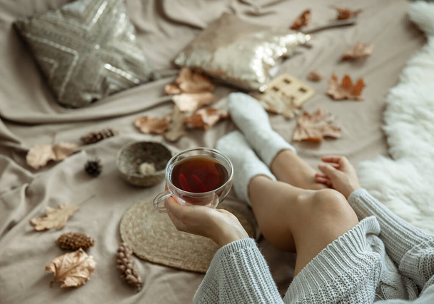 Κορίτσι σε ένα πλεκτό πουλόβερ κρατά ένα φλιτζάνι τσάι στο χέρι της, θολή φόντο φθινόπωρο, σύνθεση στο σπίτι. - Φωτογραφία, εικόνα