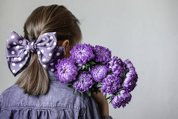 Bambina con un bel fiocco sui capelli tiene un mazzo di crisantemi blu, vista posteriore. - Foto, immagini