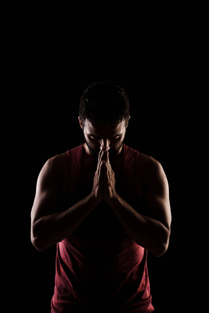Side lit muscular Caucasian man silhouette. Athlete in red shirt praying against black background.  - Photo, image