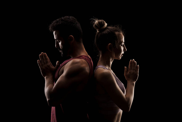 Fit couple posing together. Boy and girl praying back to back side lit silhouettes on black background. - Фото, зображення