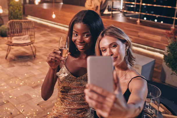 Two beautiful women in evening gowns making selfie and smiling while spending time on luxury party - Foto, afbeelding