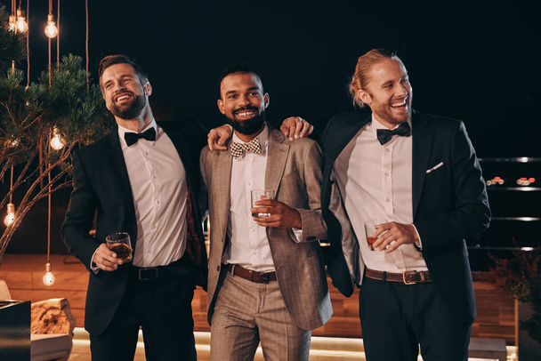 Three handsome men in suits bonding and drinking whiskey while spending time on party - Zdjęcie, obraz