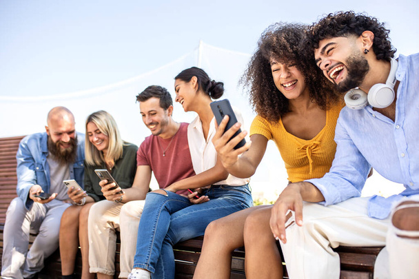 Diverse group of happy college friends using mobile phone while sitting together outdoors - Young students watching sharing content on social media - Foto, imagen