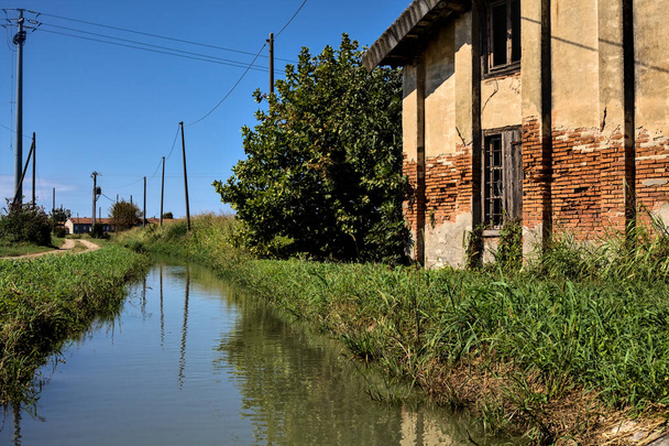 Verlaten landhuis naast een onverharde weg en een waterstroom op een heldere dag - Foto, afbeelding