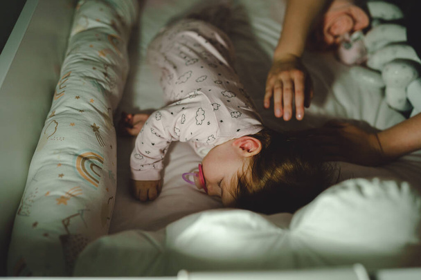 Pequeno bebê caucasiano indo dormir mãos de mulher desconhecida mãe colocando cobertor para cobrir seu pequeno filho no berço no quarto escuro à noite ou à noite - Foto, Imagem