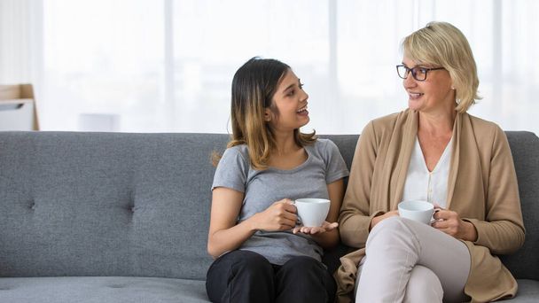 Happy middle aged woman with adopted Asian teen daughter sharing news and drinking coffee while resting together on sofa at home - Photo, Image