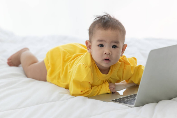 Retrato asiático bebé niño mira a cámara hacer adorable e inocente cerca con portátil en la cama en el dormitorio con luz brillante en la mañana. - Foto, imagen