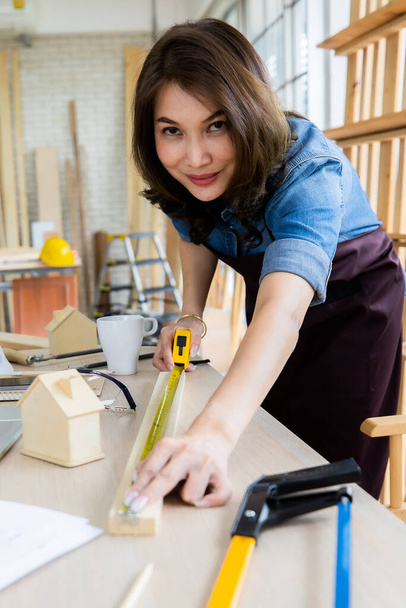 Θετική γυναίκα με ποδιά χαμογελαστή και μέτρηση ξυλείας στο τραπέζι κοντά στο laptop ενώ εργάζεται σε ελαφρά ξυλουργικά μηχανήματα - Φωτογραφία, εικόνα