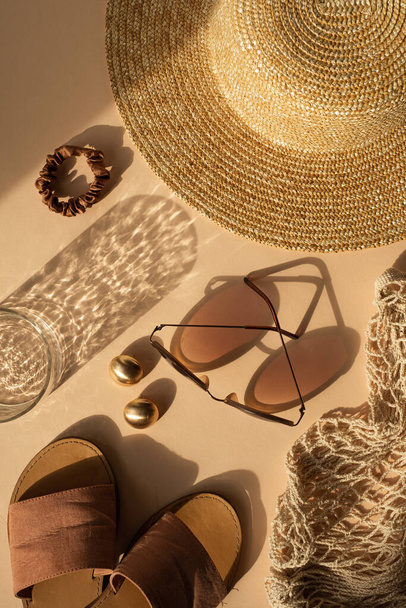 Luxury bohemian aesthetic fashion composition with crystal glass in sparkling sunlight shadows. Sunglasses, women's straw hat, string bag, gold earrings on neutral beige background. Flat lay, top view - Photo, Image