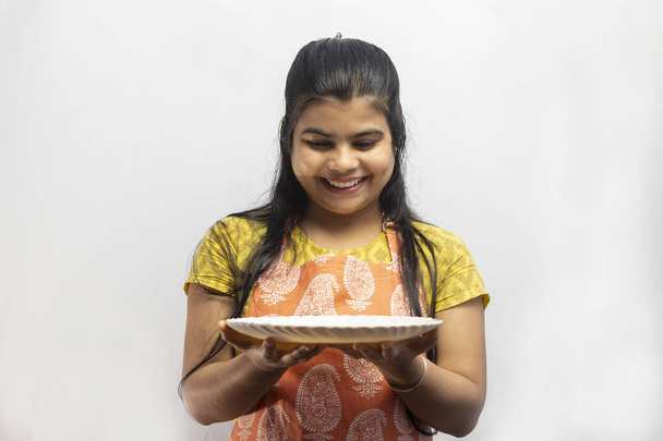 Una hermosa ama de casa india usando delantal de cocina con un plato de servir en la mano sonríe sobre fondo blanco - Foto, imagen