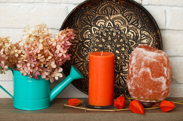 Μια σύγχρονη νεκρή φύση με αρωματικό κερί και μια λάμπα αλατιού του Νεπάλ. Φωτεινή πορτοκαλί διακόσμηση για το σπίτι και το γραφείο. Διακοσμητικό αφρικανικό πιάτο. Ποτιστήρι για λουλούδια. - Φωτογραφία, εικόνα